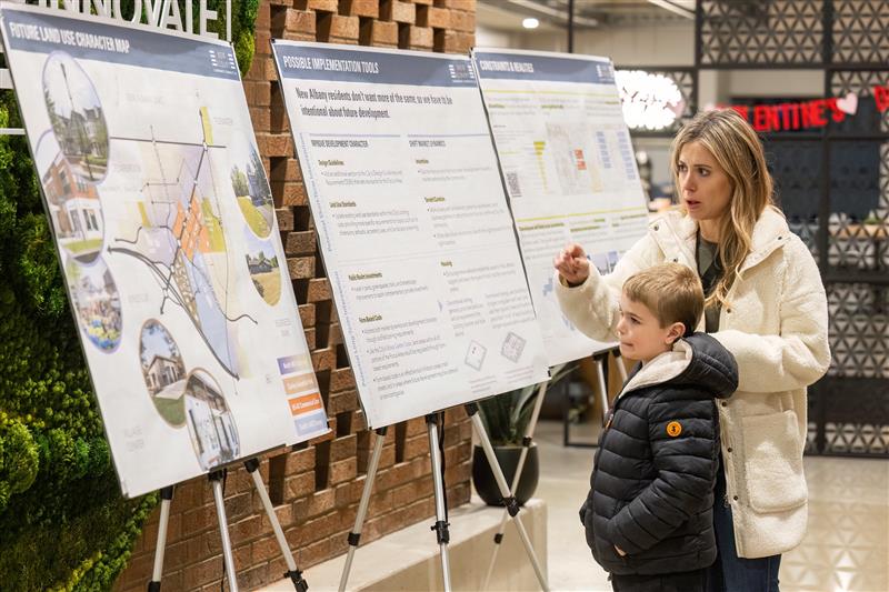 A woman in a light fleece coat points at one of three large posters with maps and text mounted on easels about future land-use plans while a young boy in a dark puffer jacket looks on in an indoor public space with a brick wall backdrop.