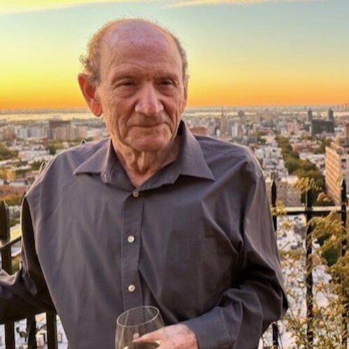 Older man in a dark button-down shirt holding a wine glass on a rooftop balcony with a city skyline and orange sunset behind him