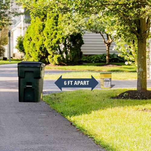 Green wheeled trash bin and a small yellow bucket set on opposite sides of a suburban driveway with a blue double-headed arrow between them labeled '6 FT APART'.