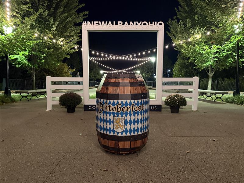 Wooden beer barrel painted with blue-and-white Bavarian diamonds and the word Oktoberfest standing in the center of a plaza at night, framed by a white photo-frame sign reading #NEWALBANYOHIO with string lights overhead, potted mums and benches in the background.