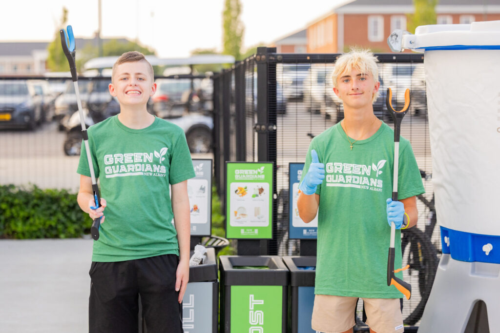 Two teen volunteers wearing green "Green Guardians New Albany" T-shirts stand outdoors in front of recycling bins holding litter pickers, one smiling broadly and the other giving a thumbs-up while wearing blue gloves.
