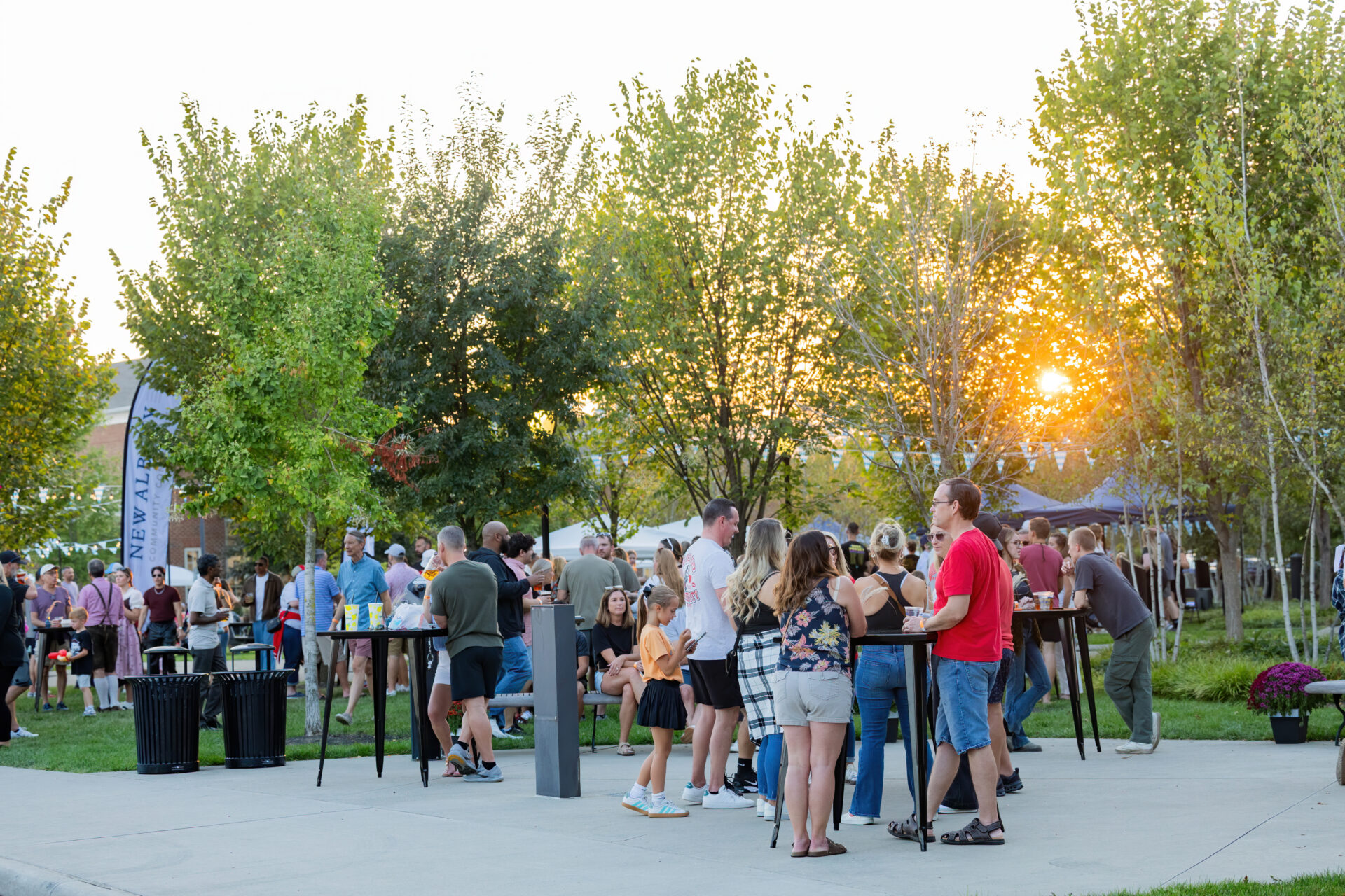 Casual crowd of adults and children gathered around tall tables and food tents in a tree-lined park at sunset, the sun glowing through the trees as people chat and hold drinks.