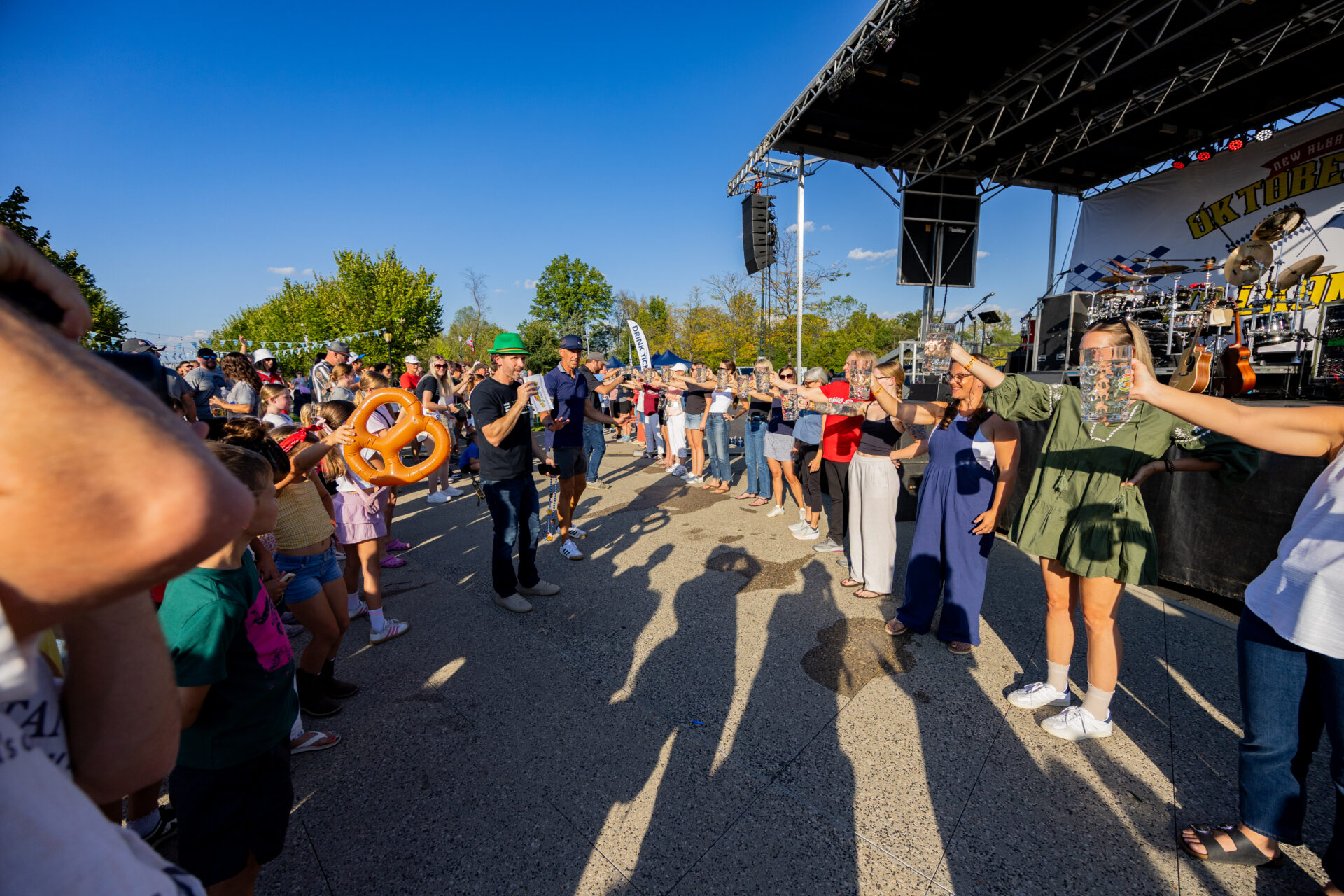 A sunny outdoor festival with a long line of people in front of a stage holding up beer steins for a group toast as an emcee in a green hat speaks into a microphone and a child nearby holds an inflatable pretzel, their long shadows stretching across the pavement.