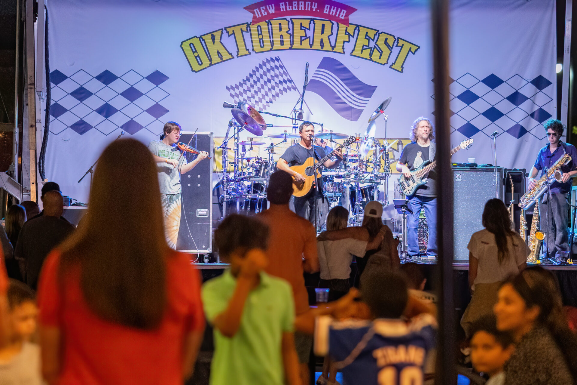 A band — including a singer with acoustic guitar, a violinist, a bassist, a saxophonist and a drummer — performs on a brightly lit stage beneath a large "Oktoberfest" banner while a crowd watches in the foreground.