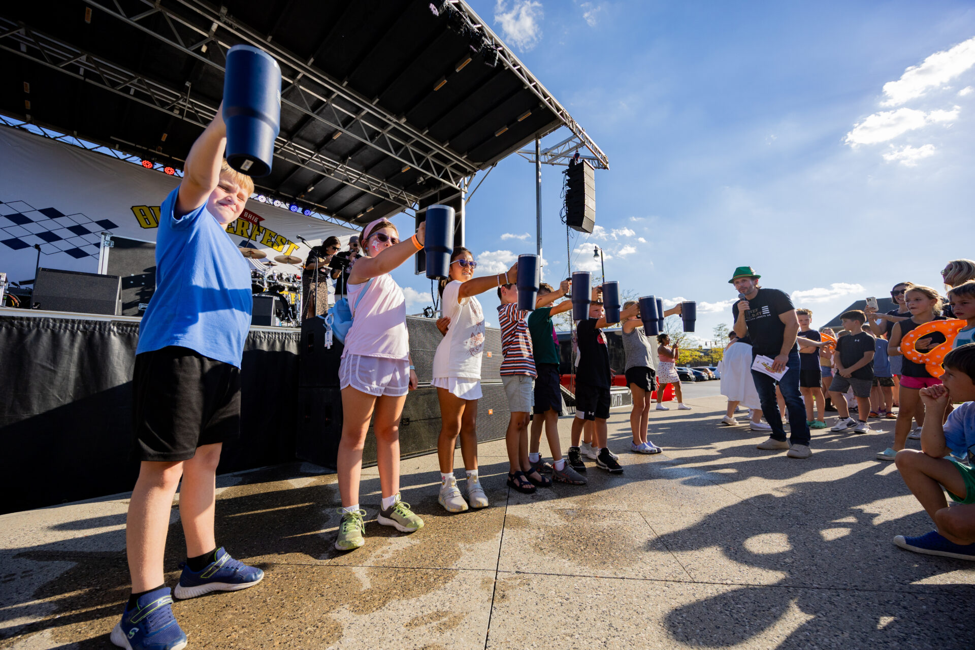 Children lined up at the front of an outdoor stage on a sunny day holding up blue tumblers for a contest while a man in a green hat with a microphone addresses them and an audience looks on.