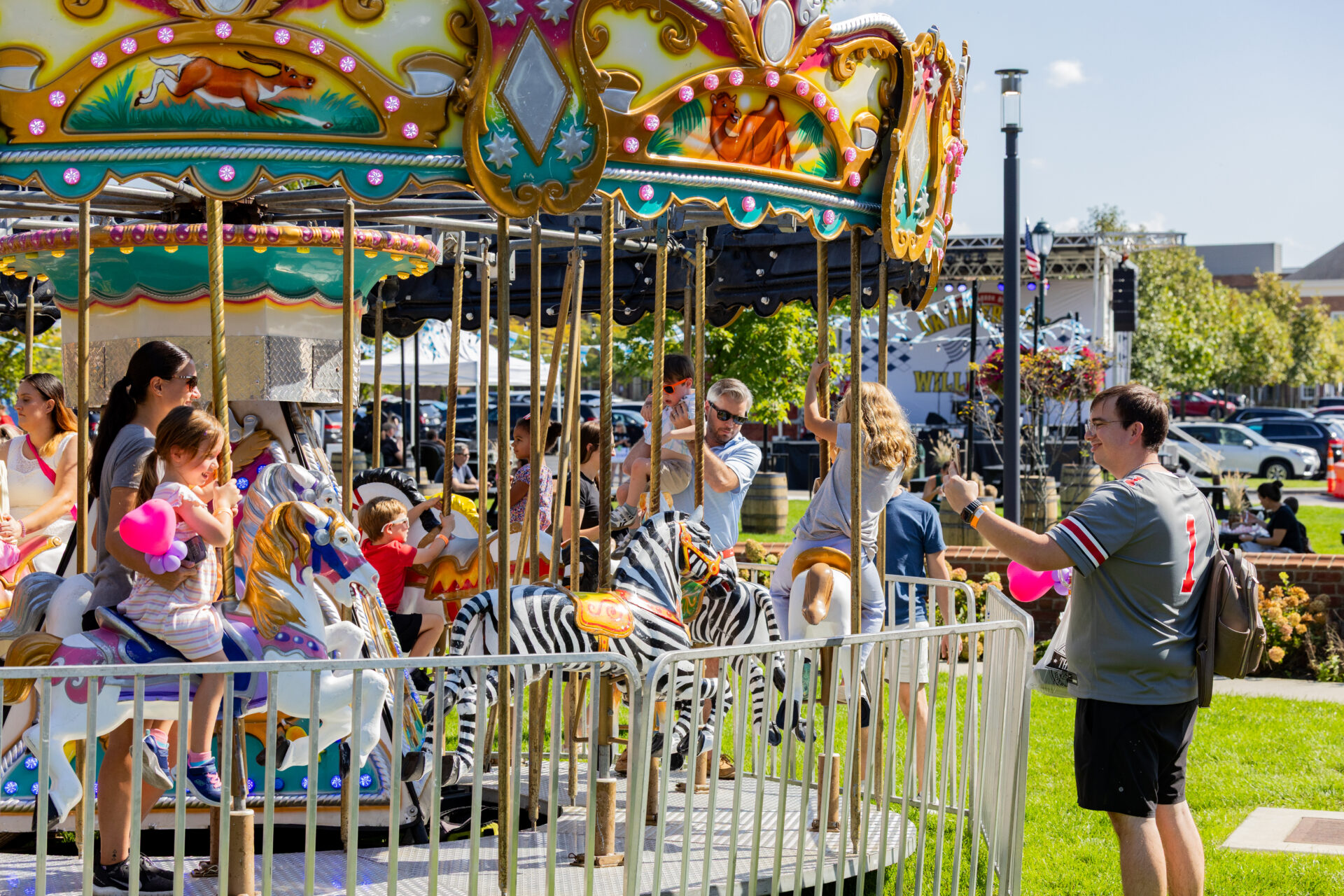 Children and parents ride a colorful vintage carousel with painted horses and a zebra while a man outside the fenced platform takes a photo on a sunny day at an outdoor fair.