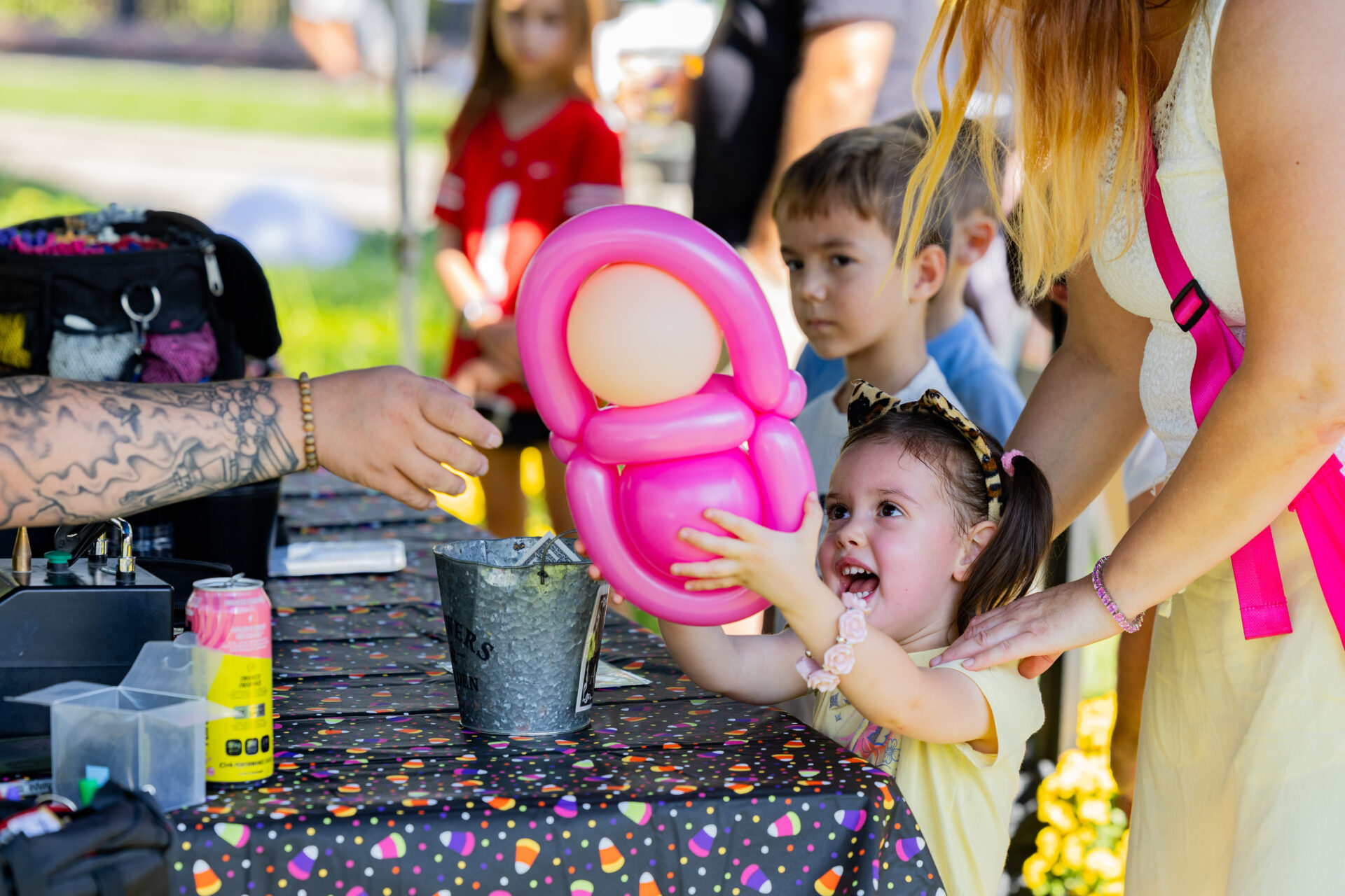 A delighted young girl with pigtails and a leopard-print headband reaches for a bright pink balloon sculpture offered across a candy-corn patterned table while a tattooed arm hands it over and an adult steadies her.