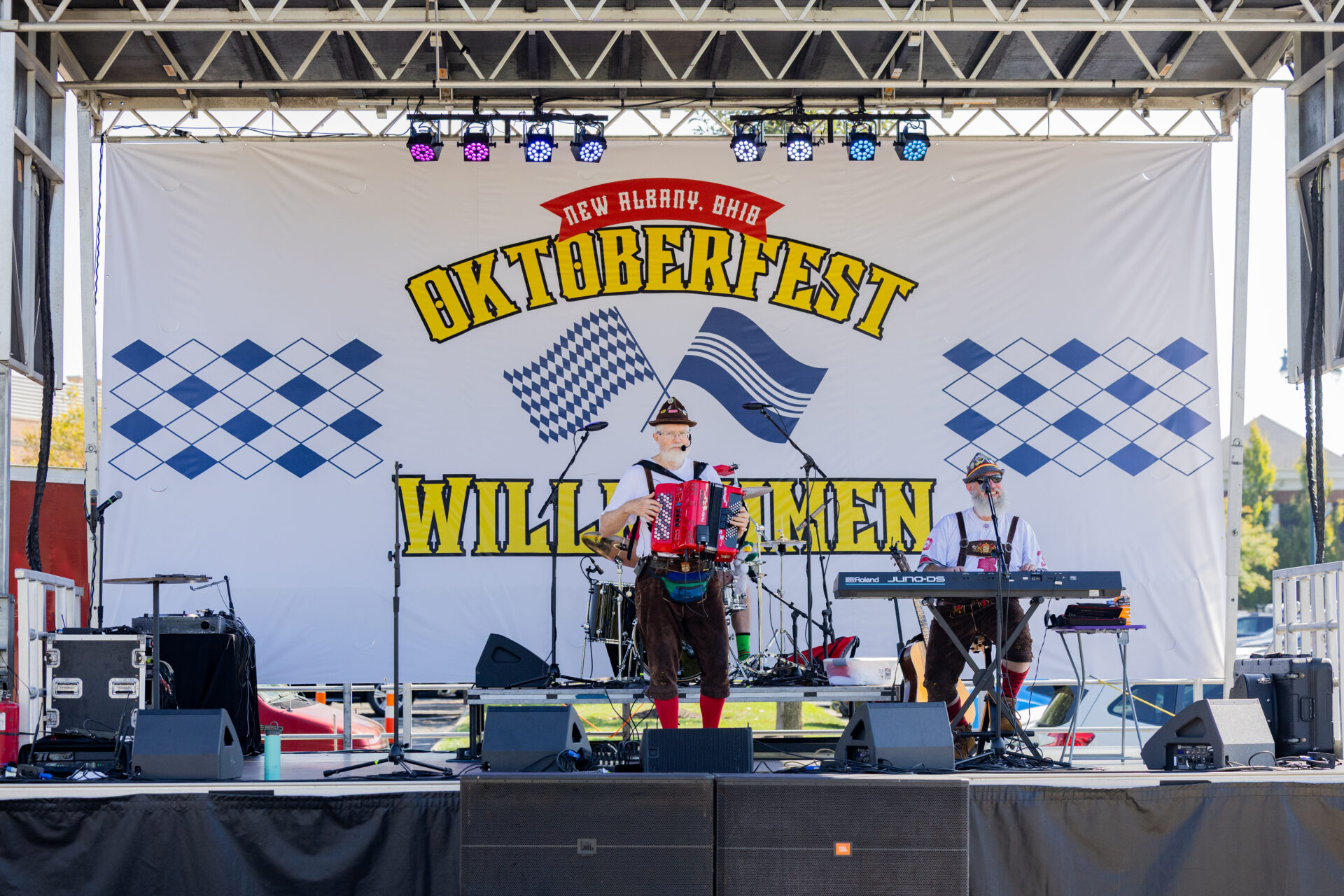 Two musicians in lederhosen perform on an outdoor stage under a large 'Oktoberfest' banner—one center playing a red accordion and the other at a keyboard—blue Bavarian diamond patterns flank the banner and stage lights hang overhead.
