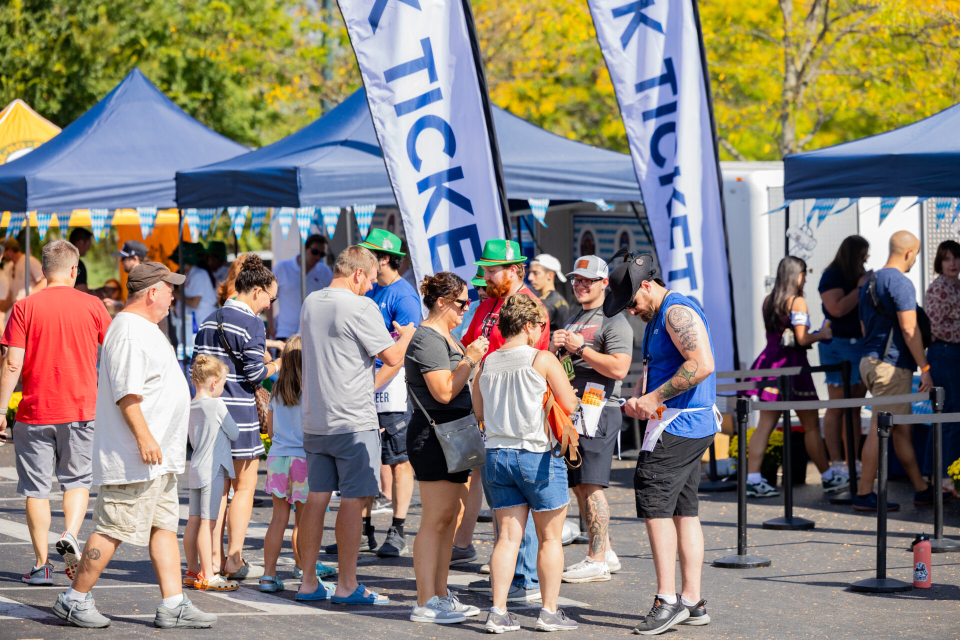 People queuing at an outdoor festival ticket area beneath blue canopy tents and tall 'TICKET' flags, including families, attendees in green hats, and a tattooed man in a black cowboy hat checking tickets.