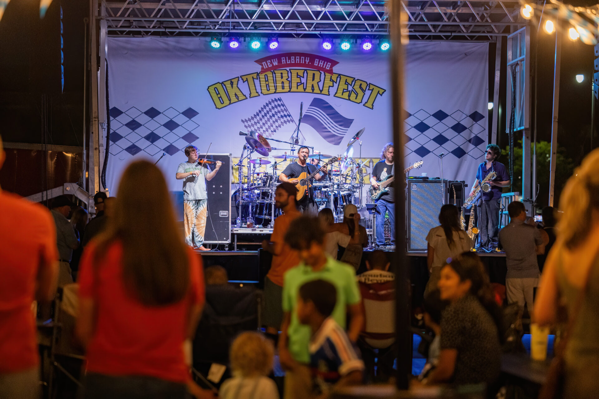 A five-member band — including a violinist, guitarist, bassist, drummer and saxophonist — performs on a brightly lit outdoor stage beneath a large 'Oktoberfest' banner as an evening crowd watches.