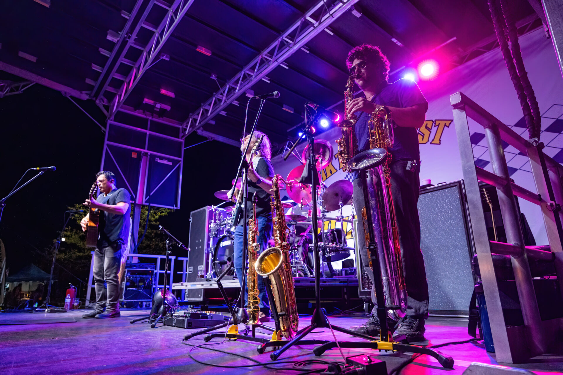 A saxophonist wearing sunglasses plays on a brightly lit outdoor stage while a guitarist and bassist perform near a drum kit, with several saxophones on stands and purple-blue concert lighting overhead.