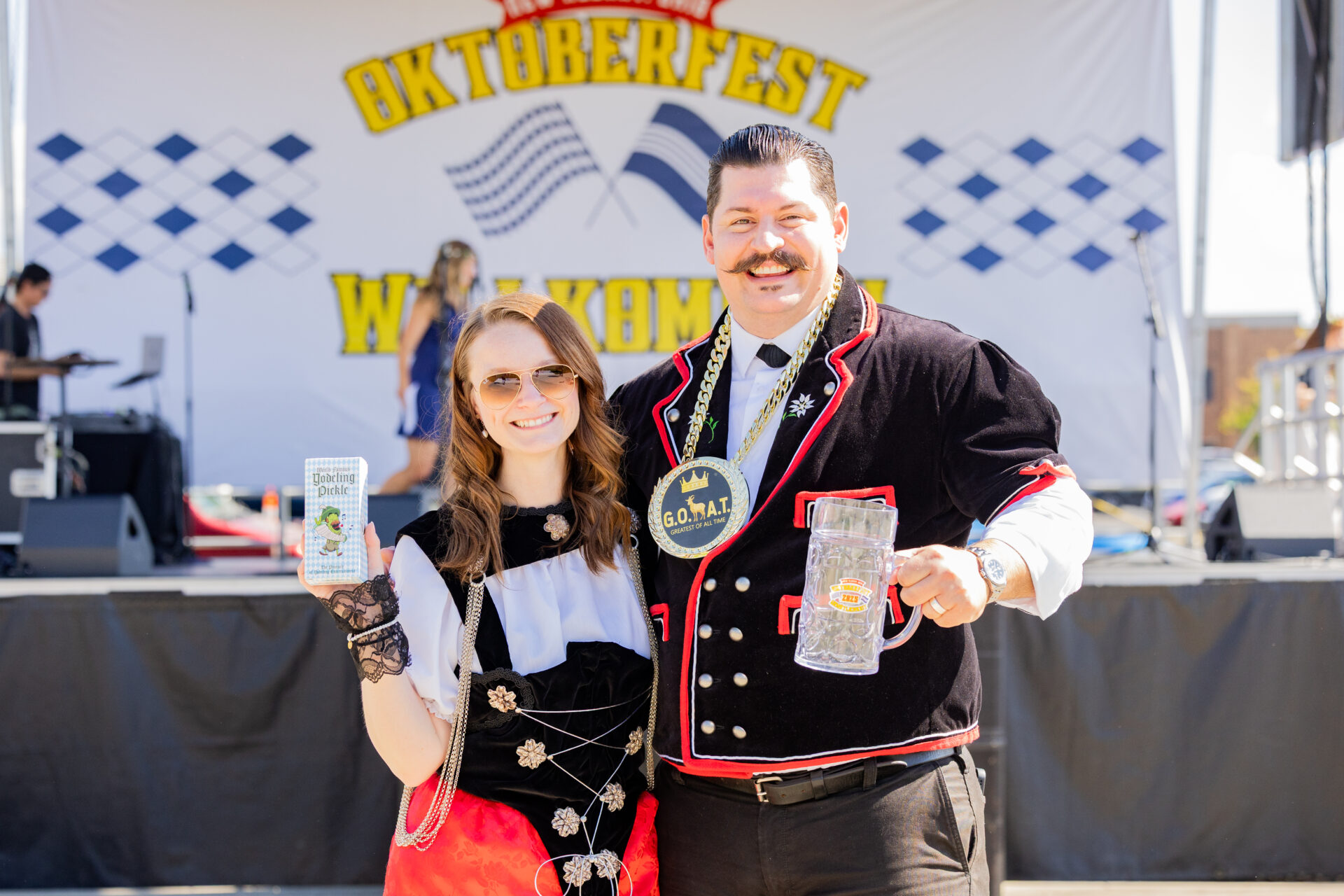 Two smiling people in traditional Oktoberfest clothing, a woman in a dirndl and sunglasses holding a packaged Yodeling Pickle and a man with a thick mustache wearing a large decorative medal and holding a clear beer stein, posed in front of a stage with an Oktoberfest banner.