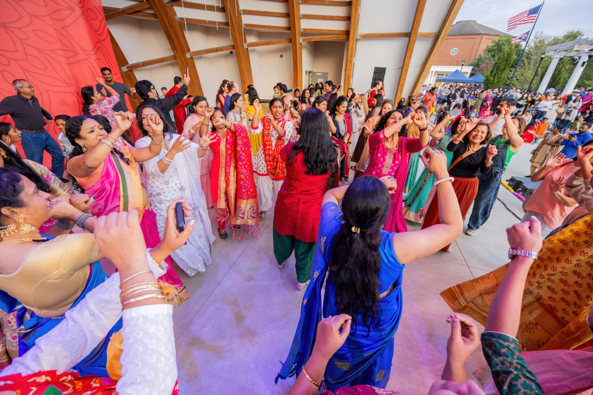 A lively outdoor dance scene showing a large group of people in colorful traditional garments—many women in saris and salwar kameez—dancing with raised arms beneath a wooden pavilion while spectators and flags stand in the background.
