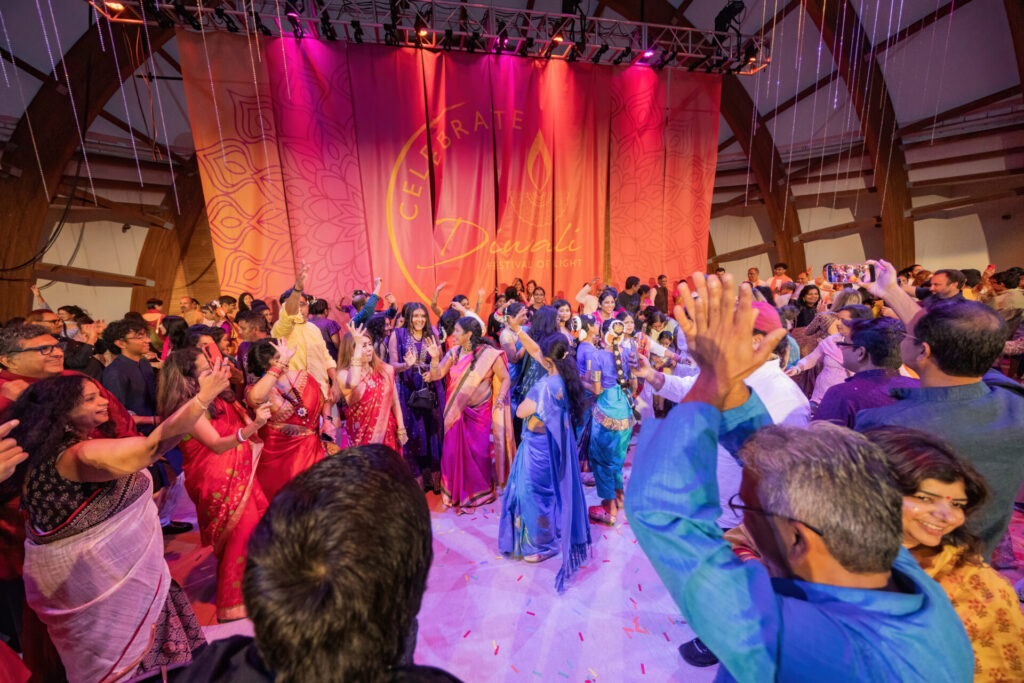 Crowd of people in colorful traditional Indian clothing dancing and celebrating in front of a large pink-orange stage backdrop that reads “Celebrate Diwali Festival of Light,” with festive lighting, hanging decorations, and confetti on the floor.