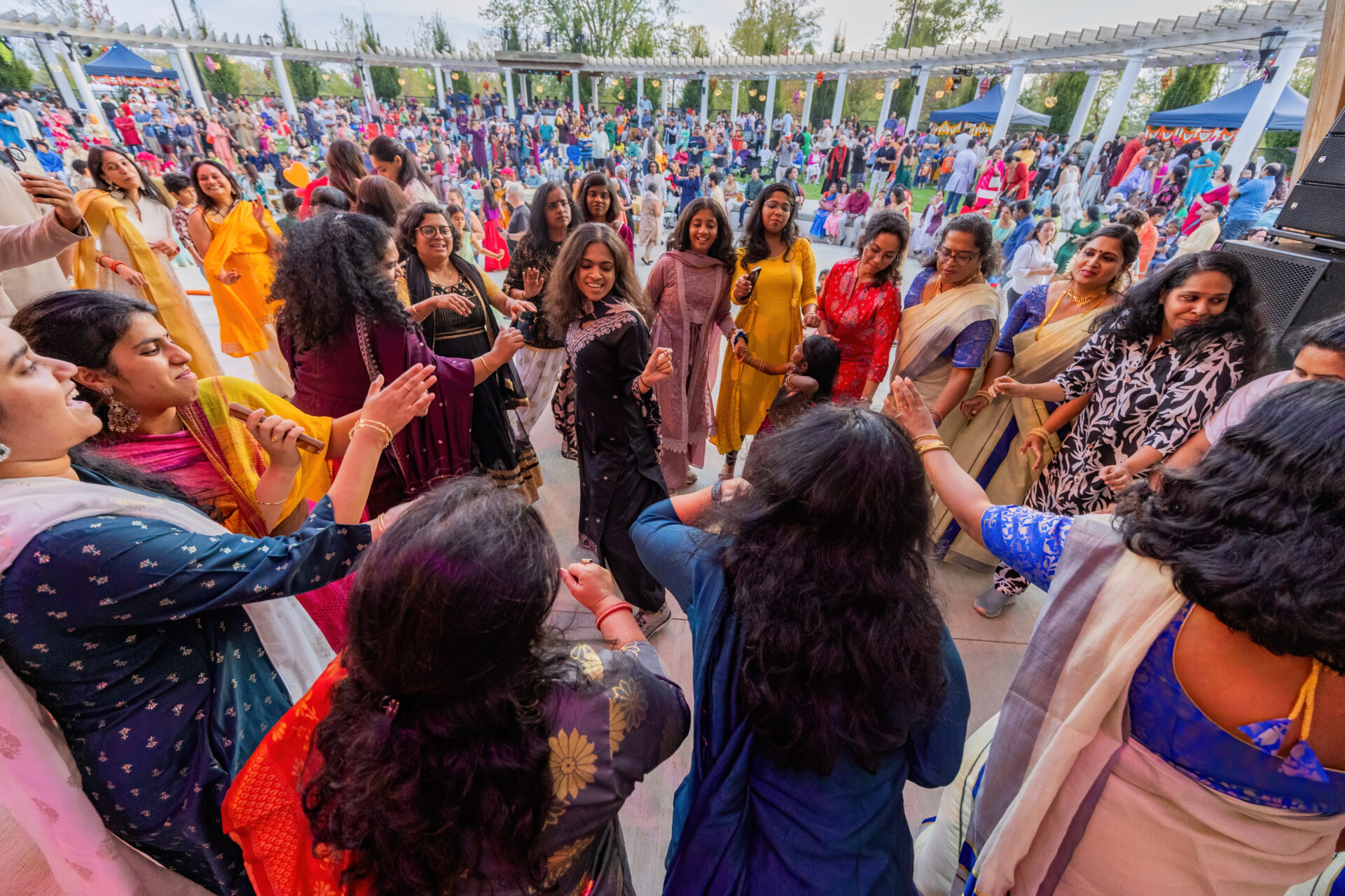 A lively circle of women in colorful traditional Indian dresses and saris dancing and smiling at an outdoor festival pavilion while a large crowd watches in the background.