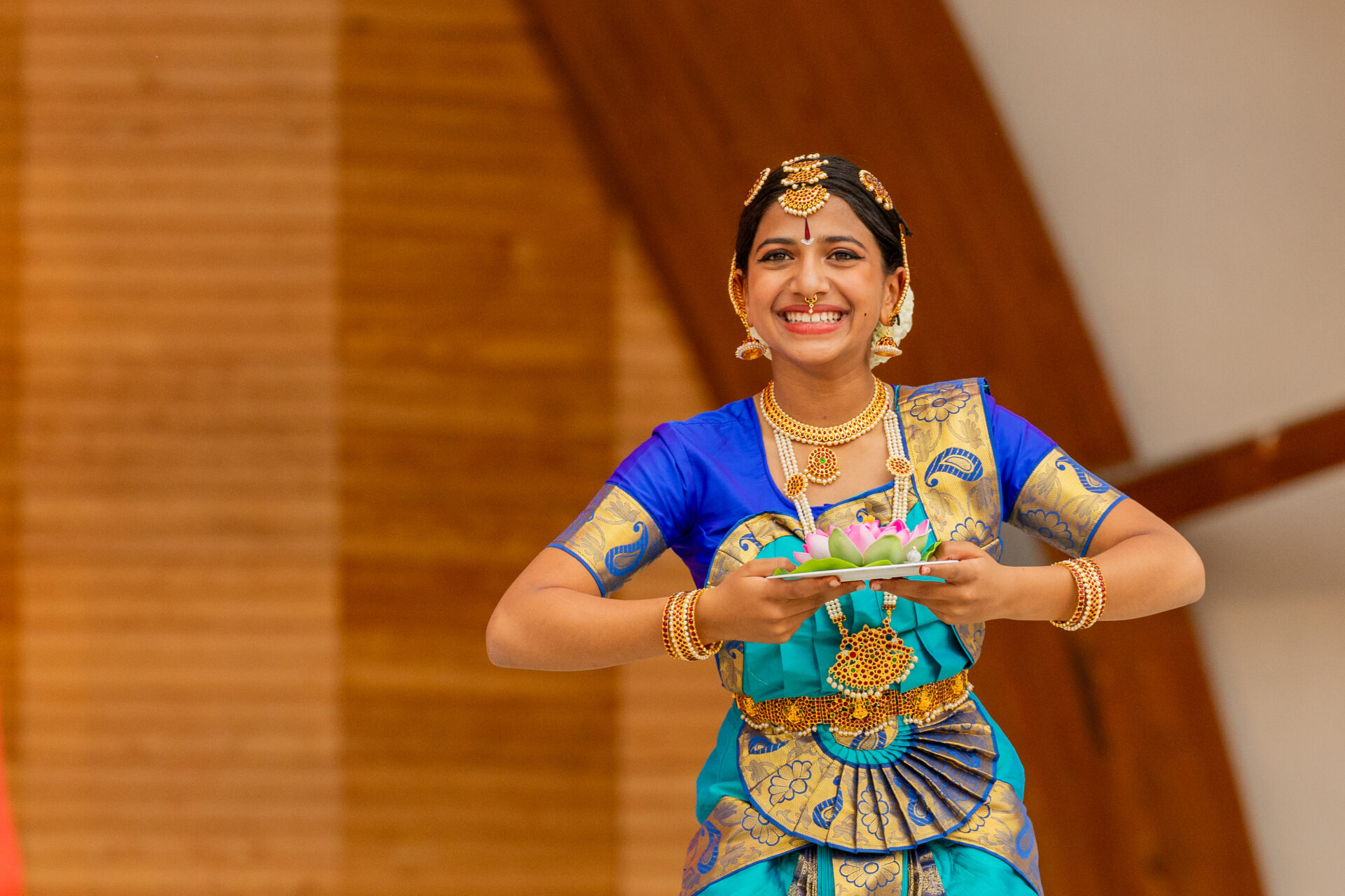 Young Bharatanatyam dancer in a blue-and-gold traditional costume and gold jewelry smiling on a wood-paneled stage while holding a small plate with a pink lotus.
