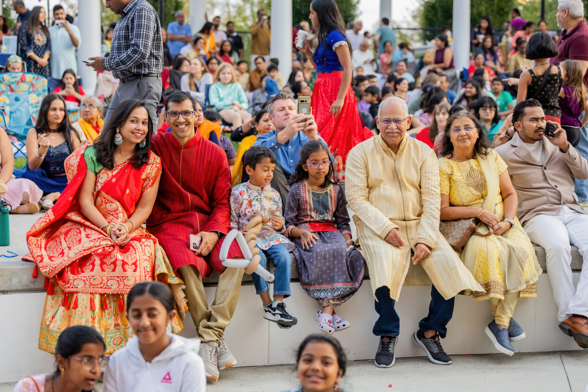 A multigenerational family in colorful traditional clothing — a woman in a red saree, a man in a red kurta, two children, and an elderly couple in pale outfits — sit on a low concrete ledge amid a large outdoor crowd at a festive event.