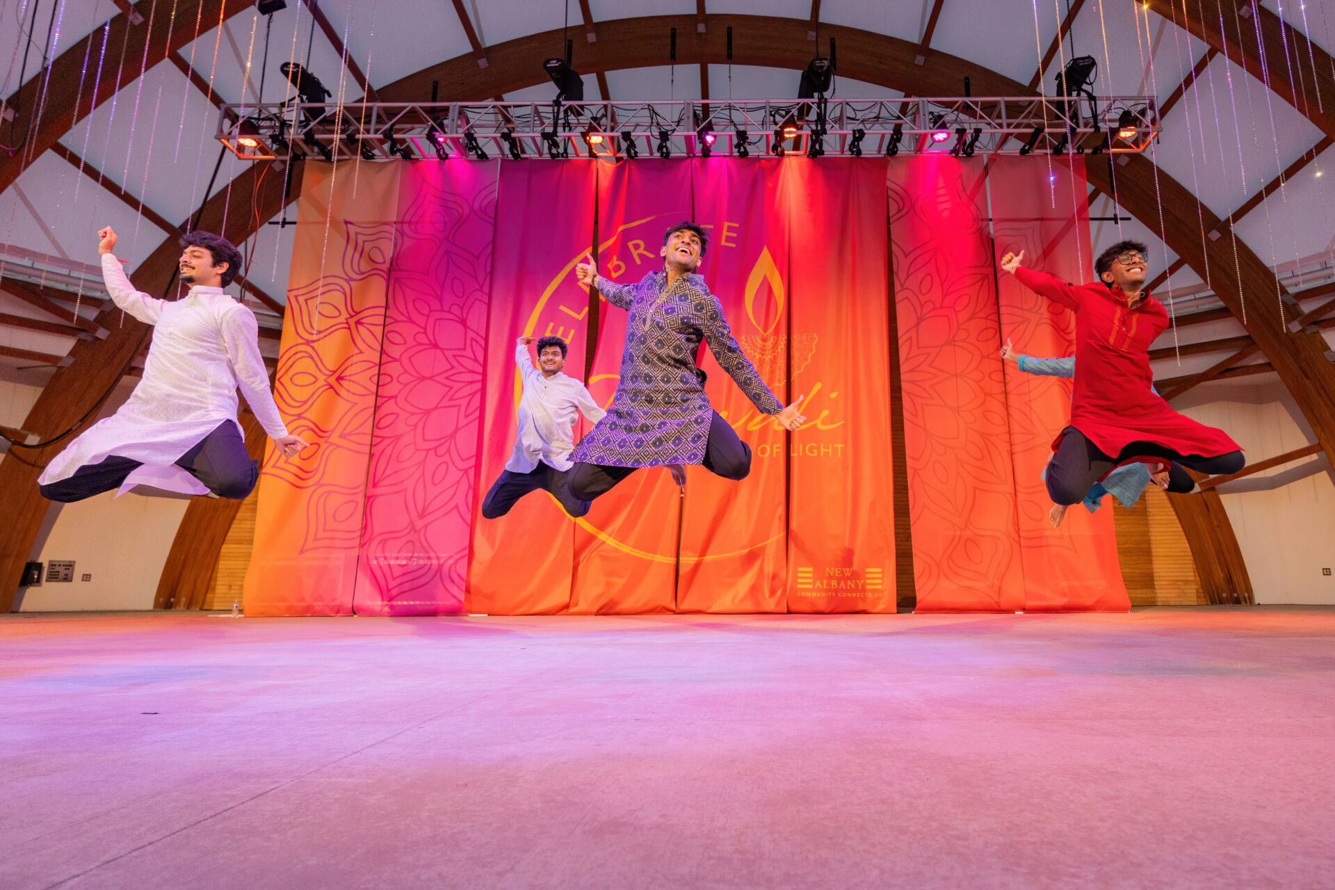 Four young men in colorful traditional kurtas leap mid-air during a lively Diwali performance on a pink‑orange lit stage with a decorative Diwali backdrop and wooden arched beams overhead.