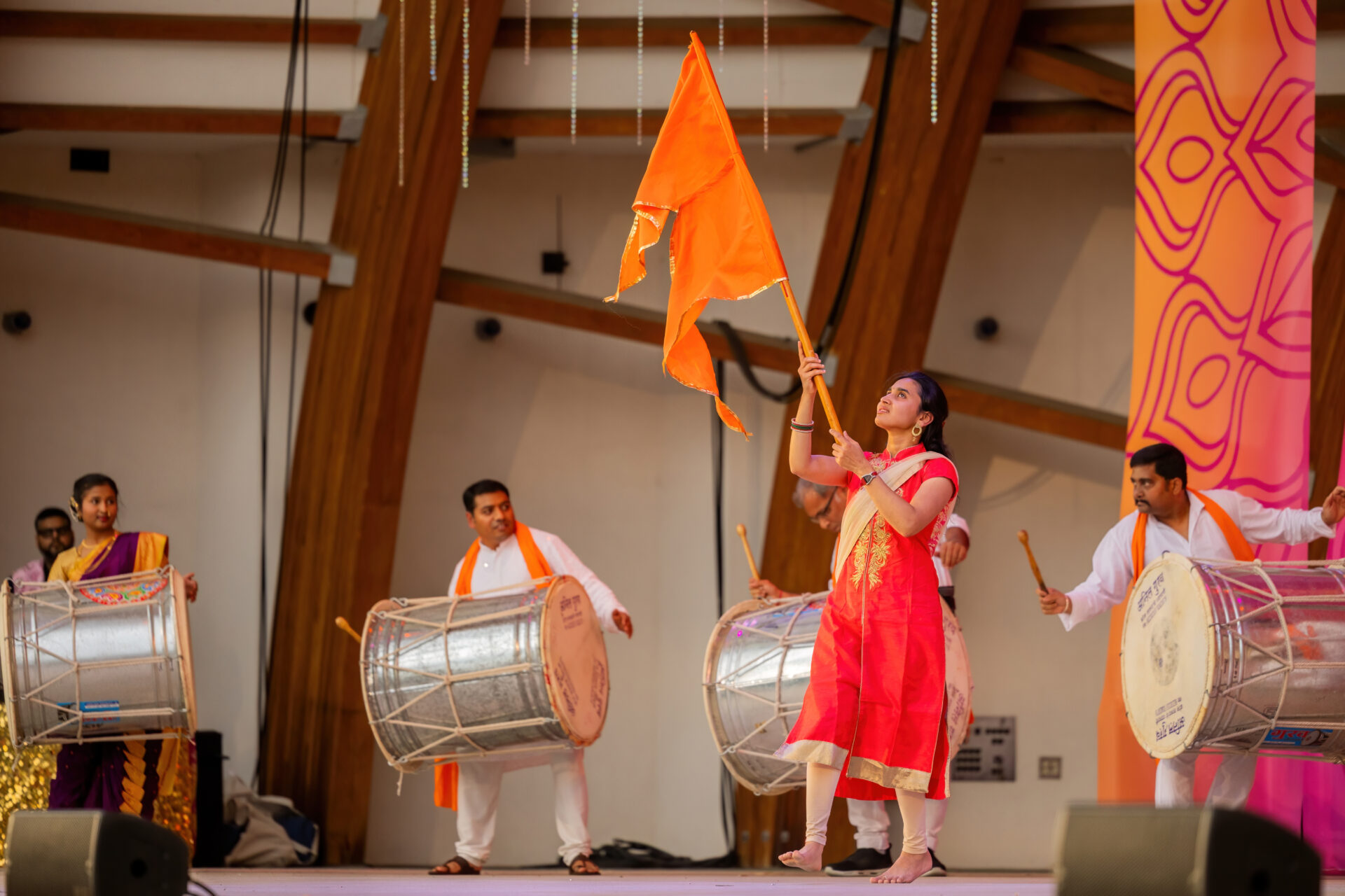 A barefoot woman in a red and gold salwar kameez waves a large orange flag onstage while four musicians in white outfits and orange scarves play large silver dhol drums beneath wooden arches and a pink-orange patterned banner.
