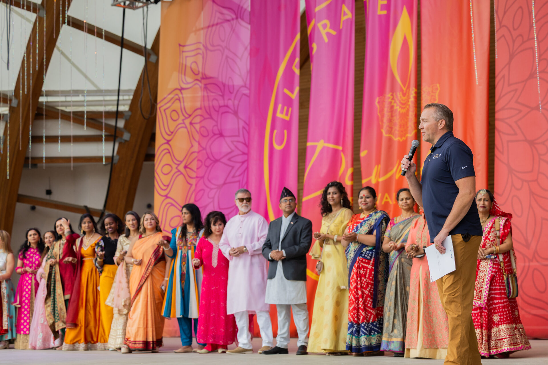 Man in a navy polo holding a microphone addresses an audience while a diverse group of men and women in colorful traditional Indian clothing stand in a line on stage in front of tall pink and orange festival banners.