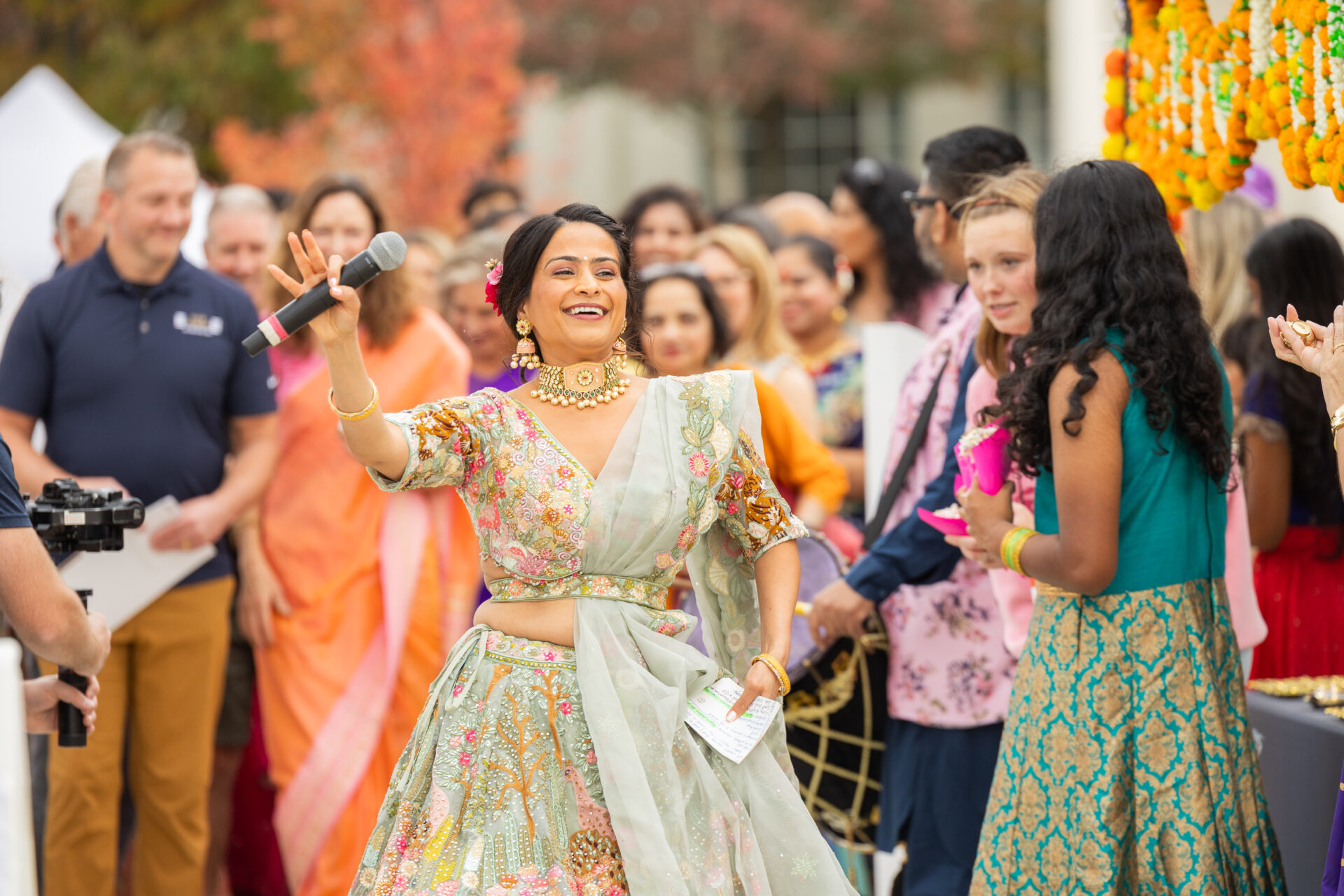 A smiling woman in a pastel green embroidered lehenga and gold jewelry, with a flower in her hair, holds a microphone and gestures energetically while addressing a crowd at a colorful outdoor celebration with marigold garlands in the background.