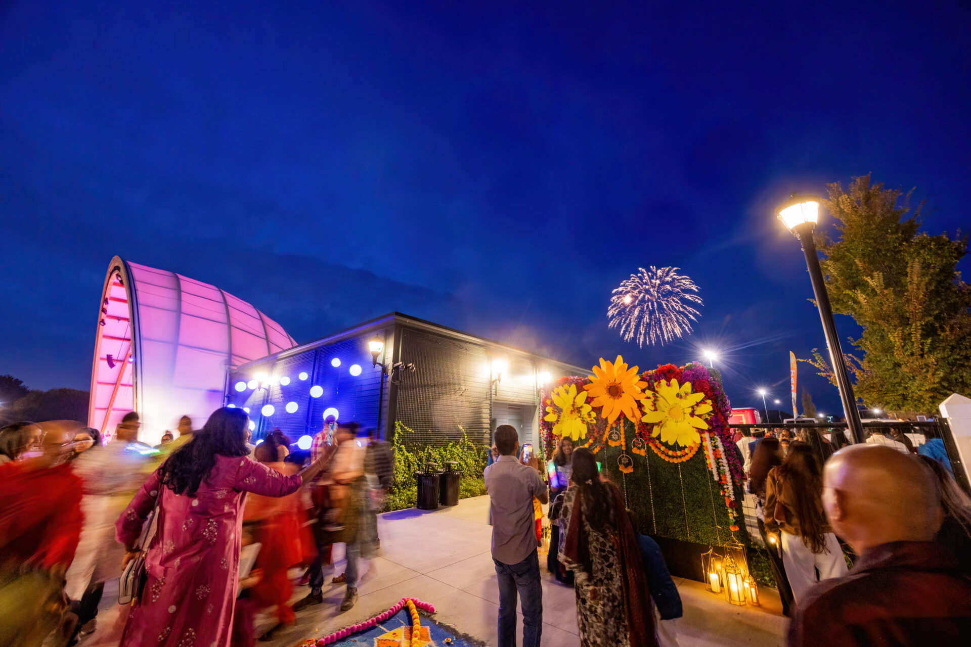 Nighttime outdoor festival with a crowd in front of a pink-lit arched stage, a large decorative floral display of sunflowers and marigold garlands to the right, lanterns and people taking photos, and fireworks bursting in the deep blue sky.