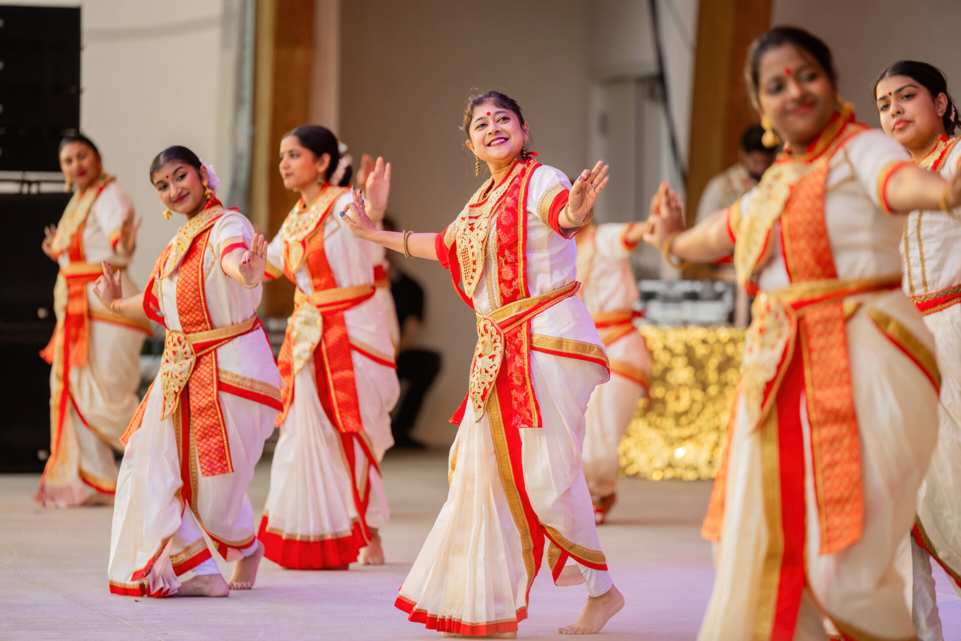 A group of women in white sarees with red-and-gold borders and matching jewelry perform a coordinated barefoot traditional dance on stage, smiling with arms extended.