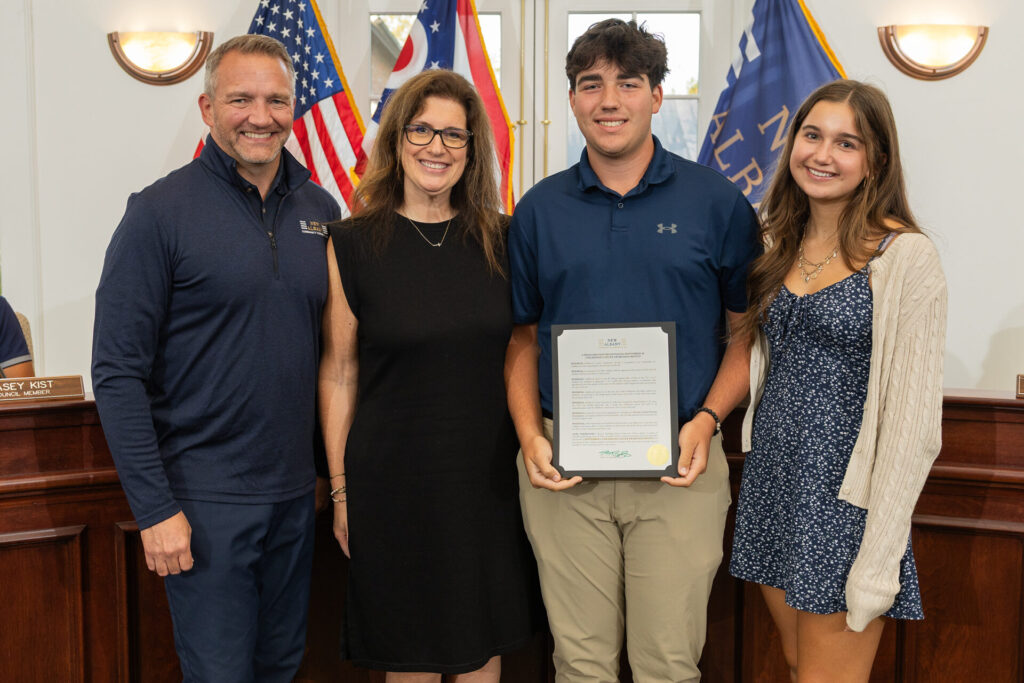 Four smiling people stand shoulder-to-shoulder in a municipal chamber with American and state flags behind them, the young man second from the right holding a framed certificate while the others pose beside him.