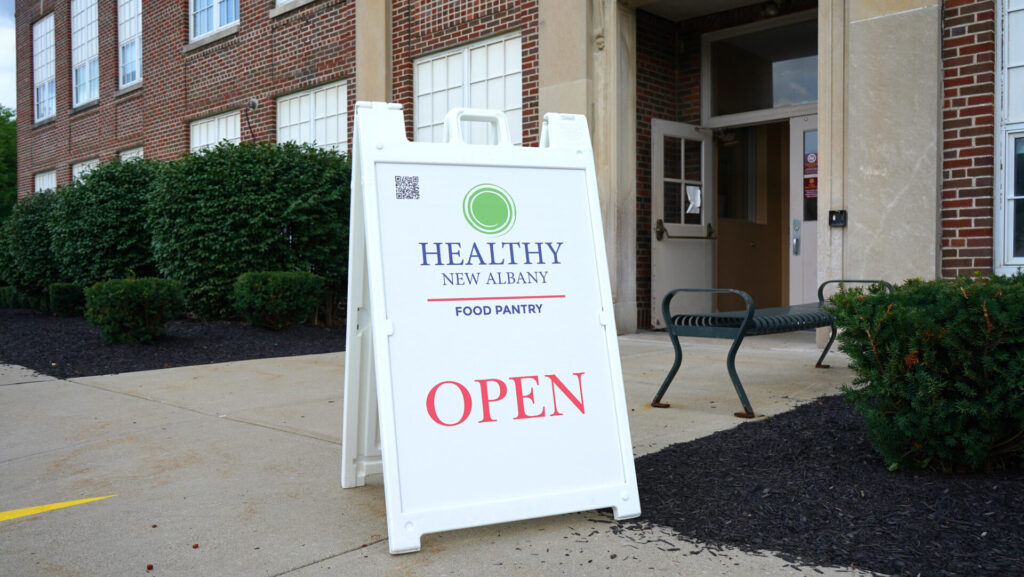 White A-frame sidewalk sign reading “Healthy New Albany Food Pantry” with a green circular logo and large red “OPEN,” placed on the sidewalk outside a brick building entrance with a bench and shrubs.