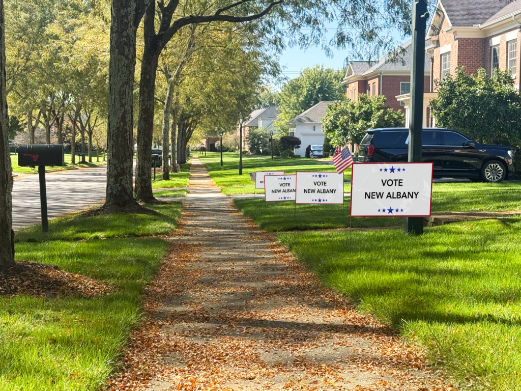 4 political signs that read "Vote New Albany" are in the grass with a sidewalk to the left.