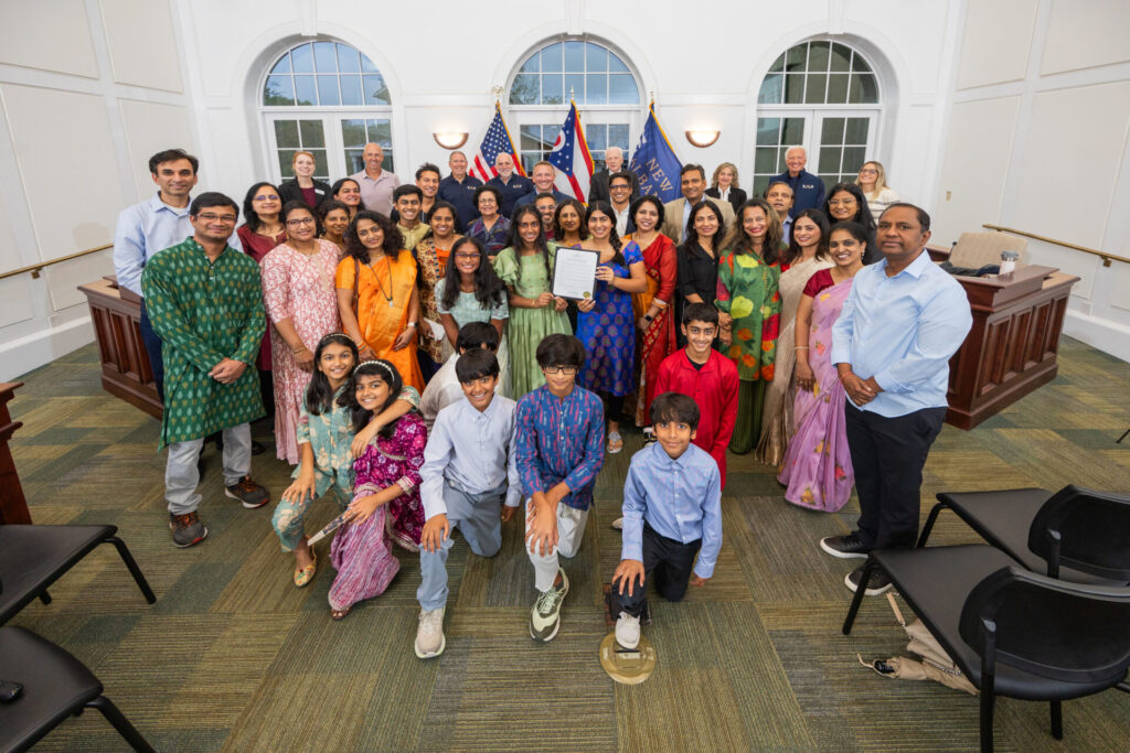 Large multi-generational group of people, many wearing colorful Indian traditional clothing, posed in a bright council chamber with flags behind them as a woman in the center holds up a framed certificate.
