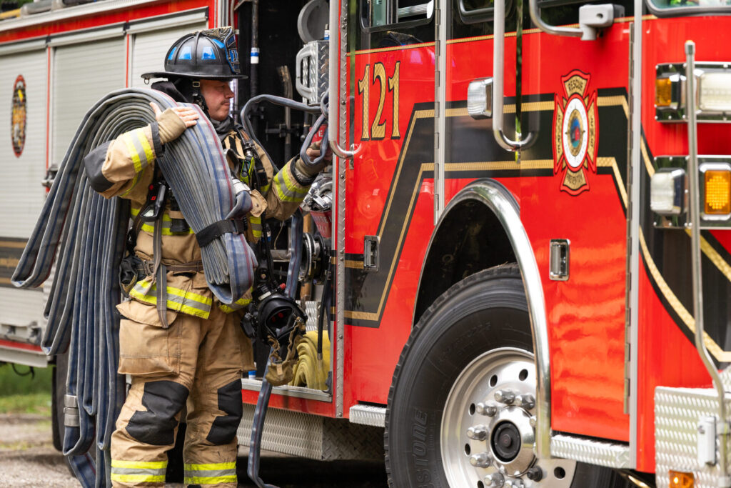 A firefighter in tan turnout gear and a black helmet carries multiple coiled fire hoses over their shoulder while reaching for a handle on a red fire engine marked 121 with reflective striping.