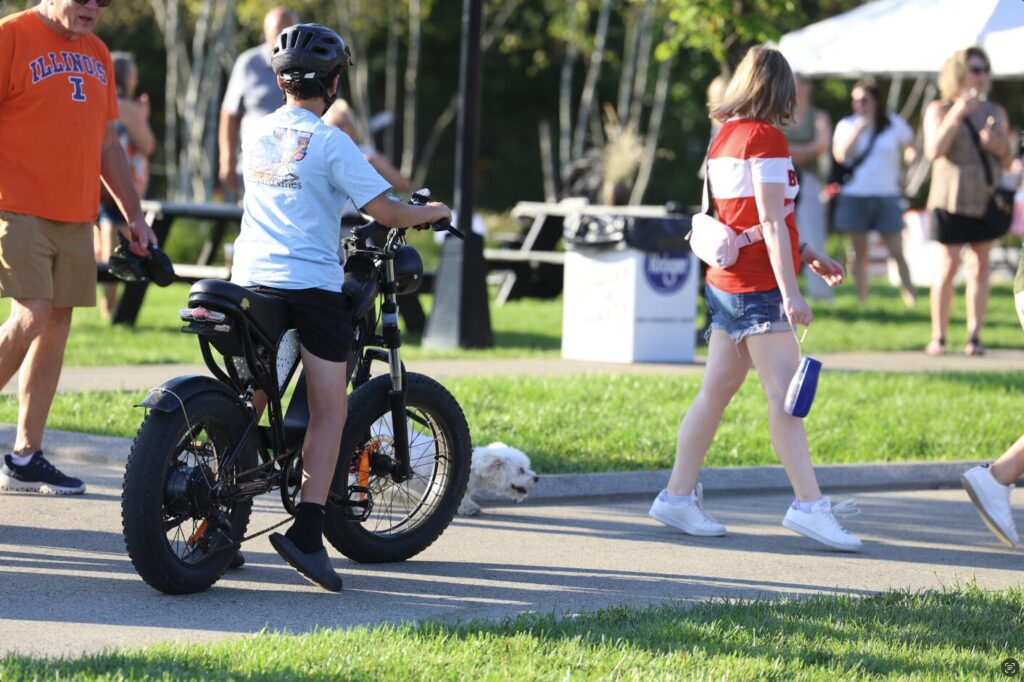 A helmeted young rider in a light-blue T-shirt sits on a black fat-tire electric bike on a park path as pedestrians, including a woman in a red-and-white shirt carrying a blue bottle and a small white dog near the pavement, pass by on a sunny day.