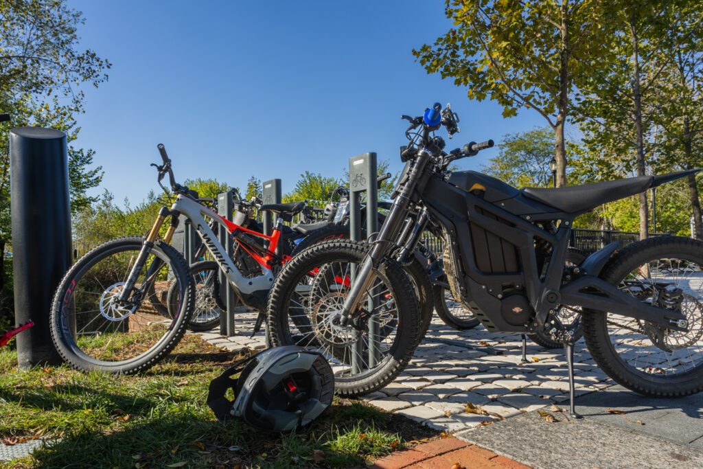 Parked mountain and electric bikes locked to a rack on a cobblestone path under trees and a blue sky, with a black electric dirt bike in the foreground and a helmet lying on the grass.