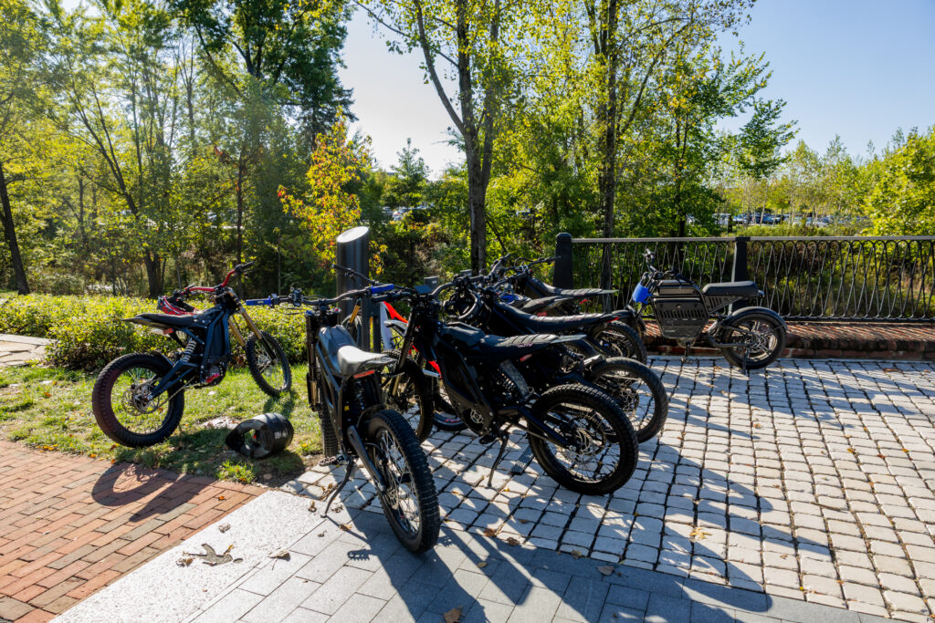 Several parked electric dirt bikes and a small electric scooter lined up on cobblestone and brick pavement beside a metal railing in a sunlit, tree-filled park, with a helmet on the grass nearby.
