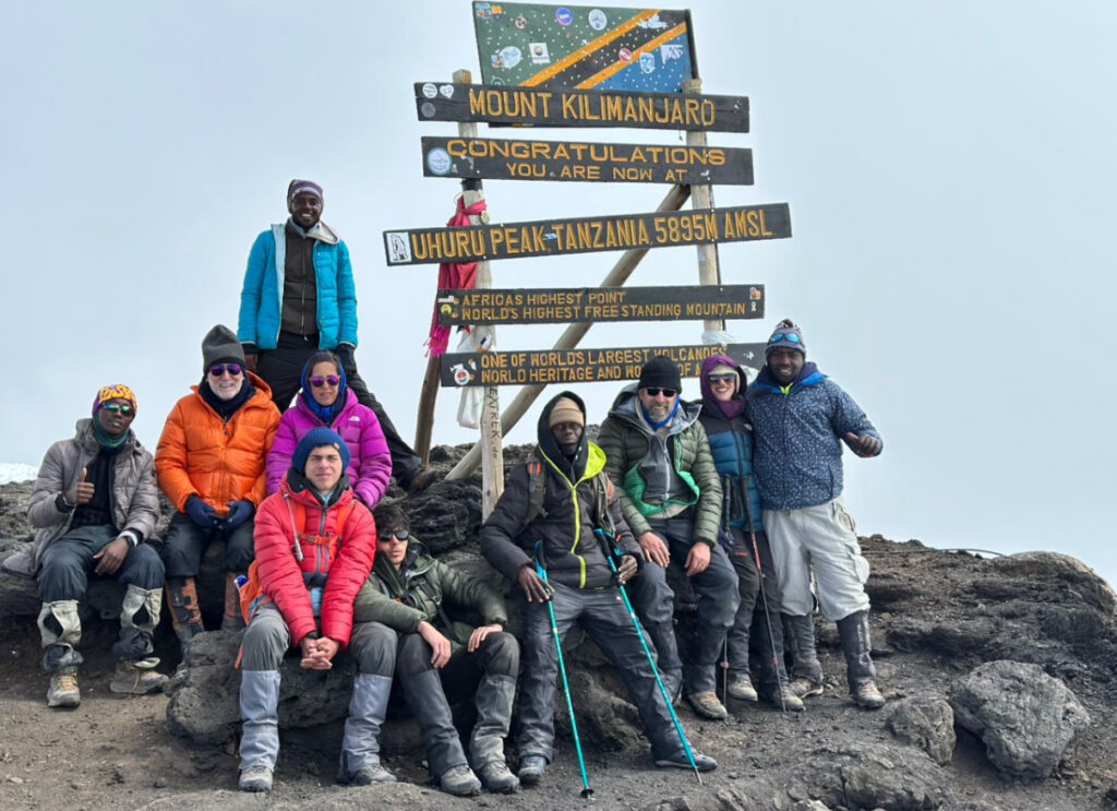 A group of climbers and guides bundled in colorful down jackets and hats pose with trekking poles around the wooden Uhuru Peak sign on Mount Kilimanjaro marking 5,895 m, standing on rocky ground under an overcast sky.