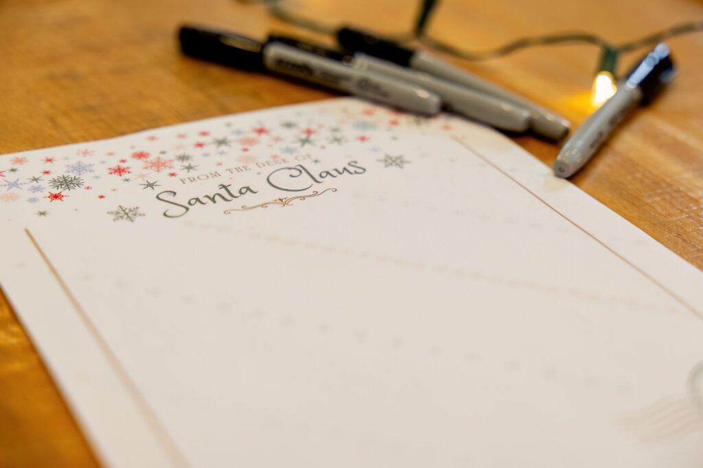 Blank "From the desk of Santa Claus" stationery decorated with multicolored snowflakes resting on a wooden table, with gray Sharpie markers and a lit string of Christmas lights nearby.