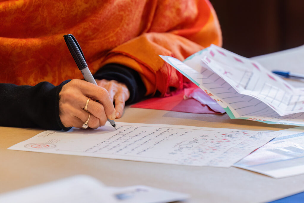 Close-up of a person wearing an orange shawl writing a handwritten holiday card with a black marker, showing ring-adorned hands and a table scattered with envelopes and folded cards.