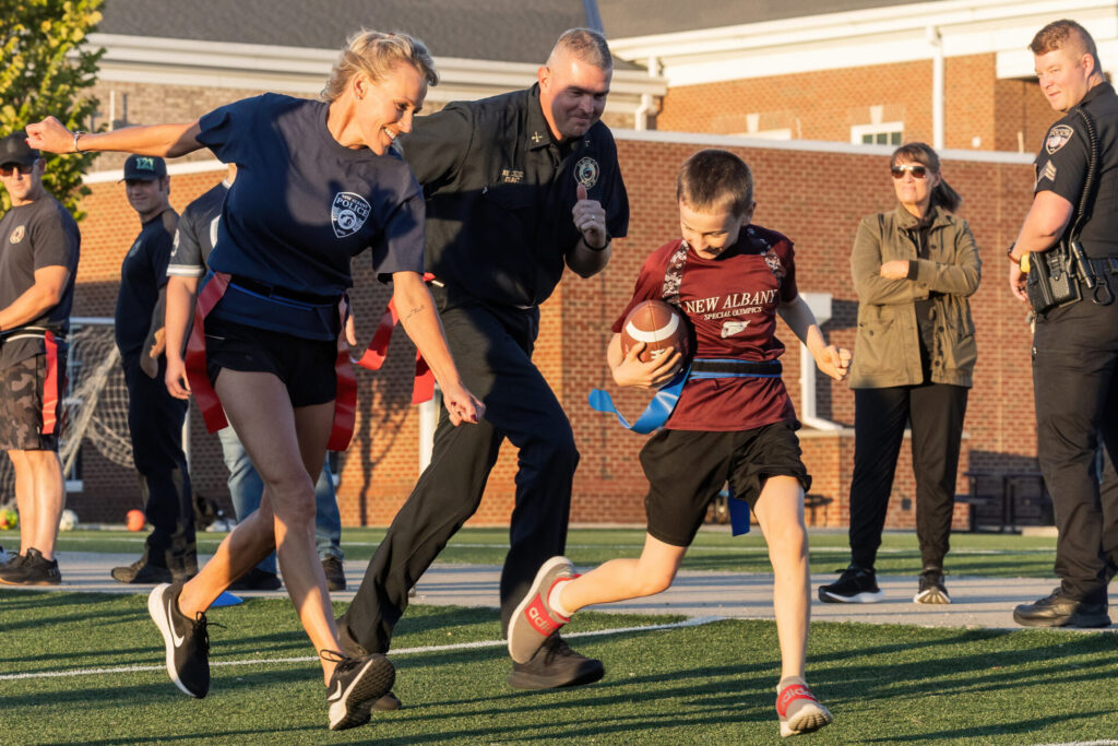 A young boy in a maroon "New Albany Special Olympics" shirt dashes with a football while two first responders reach for the blue and red flag belts around his waist on a sunlit artificial turf field in front of a brick school building.