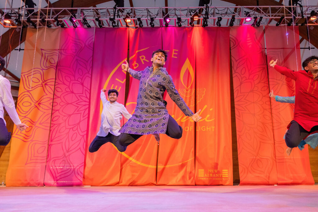 Four male dancers leap in mid-air on a brightly lit stage, wearing colorful traditional kurtas against a pink-orange backdrop decorated with mandala patterns, a Diwali flame logo, and small "New Albany" text.