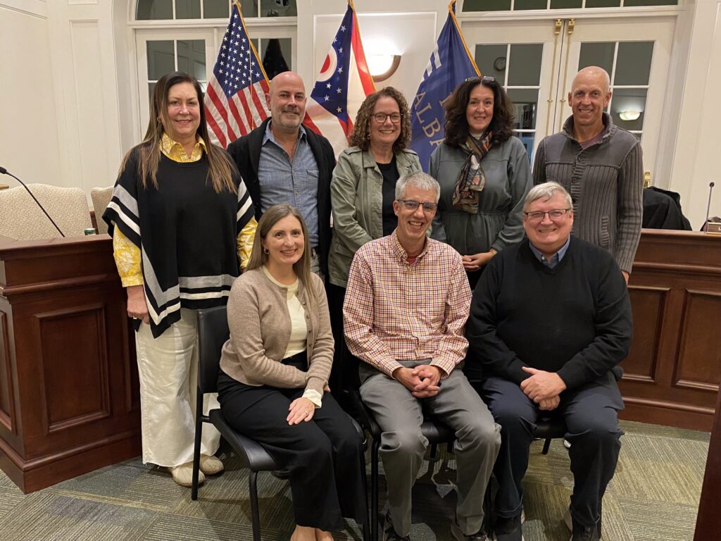 Eight adults—three seated in front and five standing behind—smile and pose in a wood-paneled meeting room with American, Ohio, and municipal flags visible behind them.