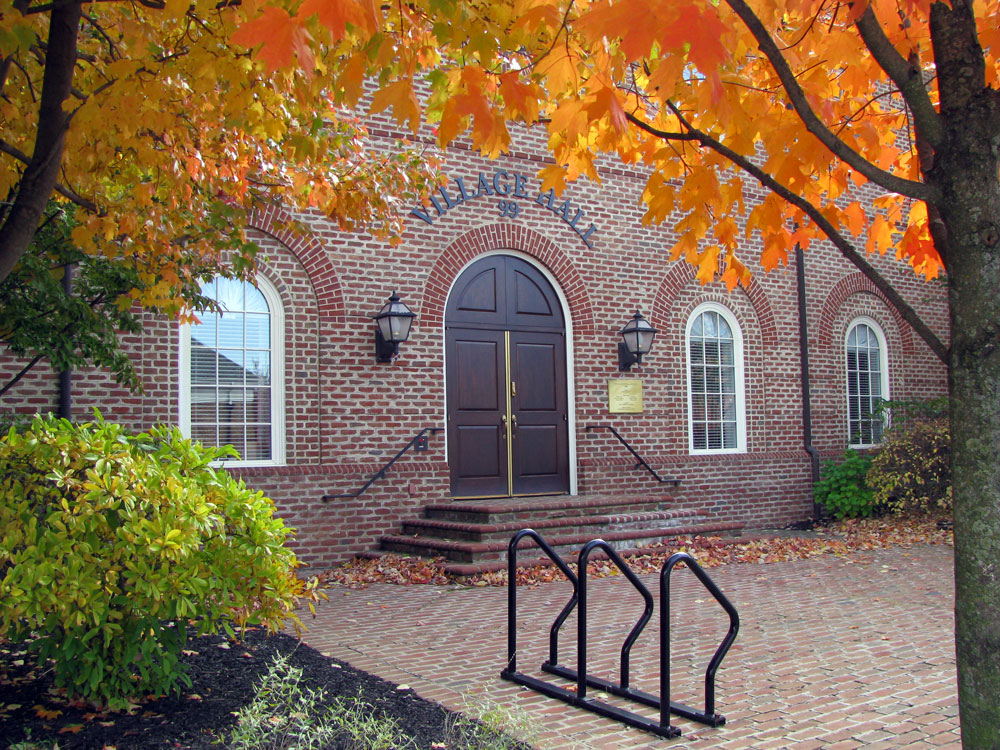 Brick village hall with arched double wooden doors and three arched windows, framed by bright orange autumn leaves and a tree, with a bike rack on the brick courtyard and steps to the entrance.