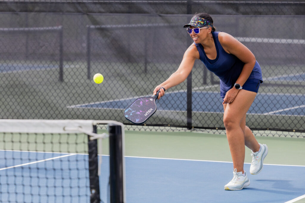 Woman in a navy tank top, visor and sunglasses leans forward on an outdoor court, smiling as she reaches with a colorful pickleball paddle to hit a yellow ball over the net, with a chain-link fence in the background.