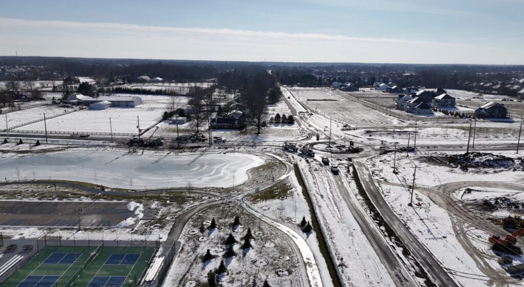 Aerial view of a snowy suburban area with a frozen pond, winding roads, tennis courts and houses under construction