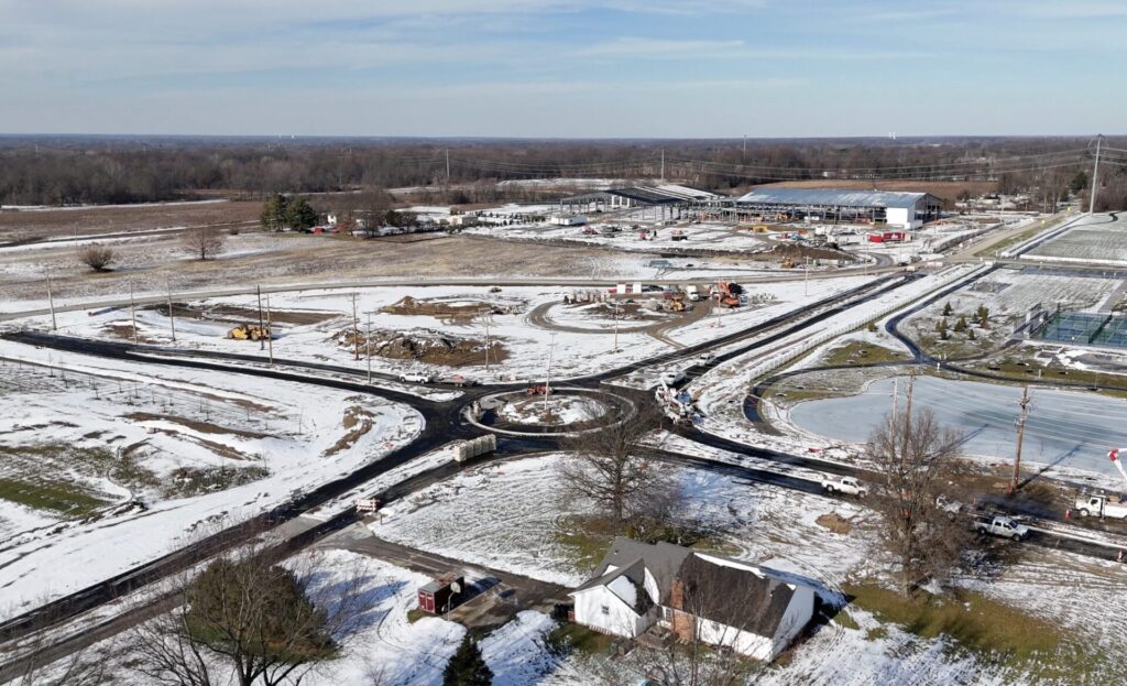 Aerial view of a snowy construction site with a newly paved roundabout, roads, construction equipment, and a farmhouse.