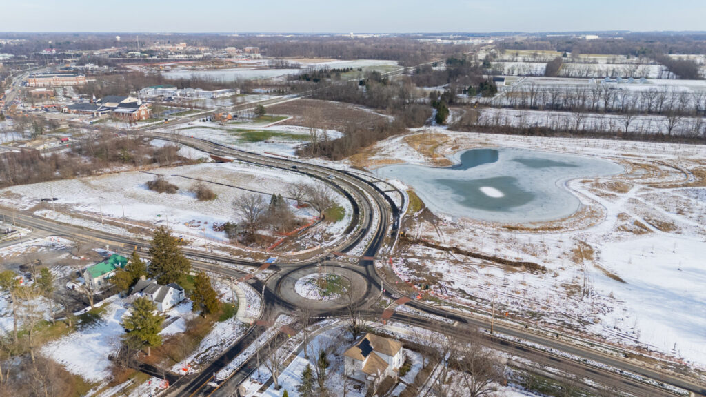 Aerial view of snow-dusted fields with a circular roundabout and curved roads beside a partially frozen pond