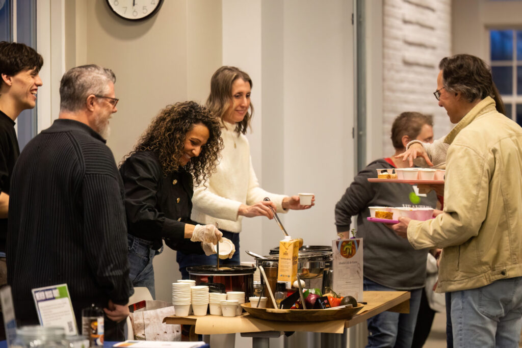 People gathered at a buffet table as a woman with curly hair ladles soup into cups while others wait with trays