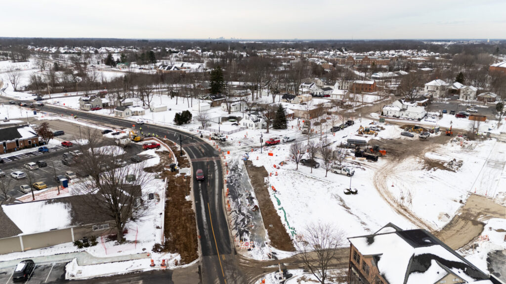 Aerial view of a snowy suburban road construction site with orange traffic cones, work trucks, parking lots, and nearby houses.