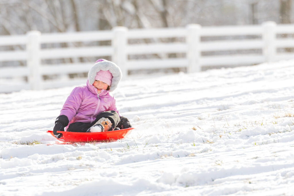 Child in a pink fur‑hooded jacket riding a red sled down a snowy hill with a white fence in the background.