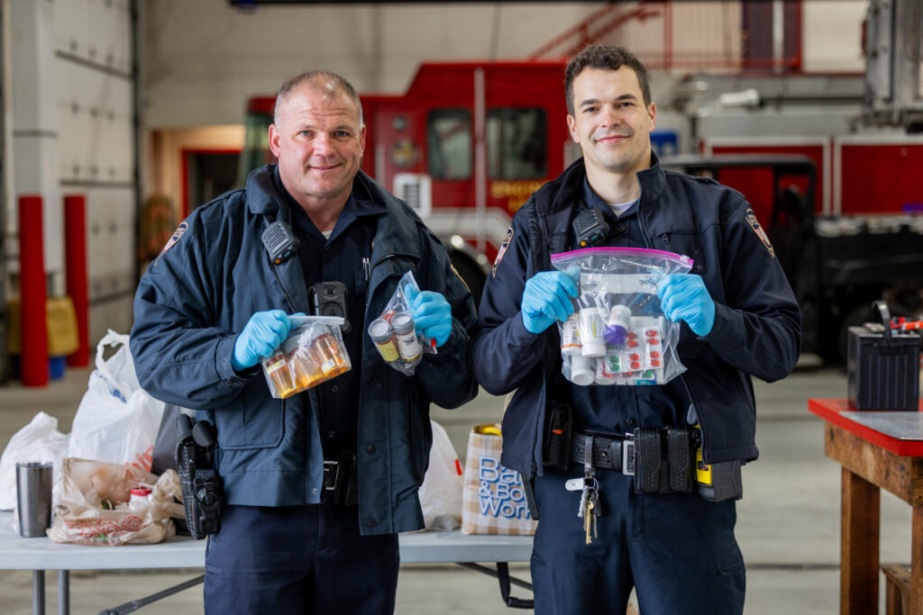 Two uniformed emergency responders at a fire station holding clear bags of prescription bottles and pills with blue gloves.